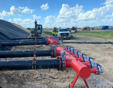 Large black pipes being installed or maintained at a construction site, with construction workers and equipment in the background under a partly cloudy sky.