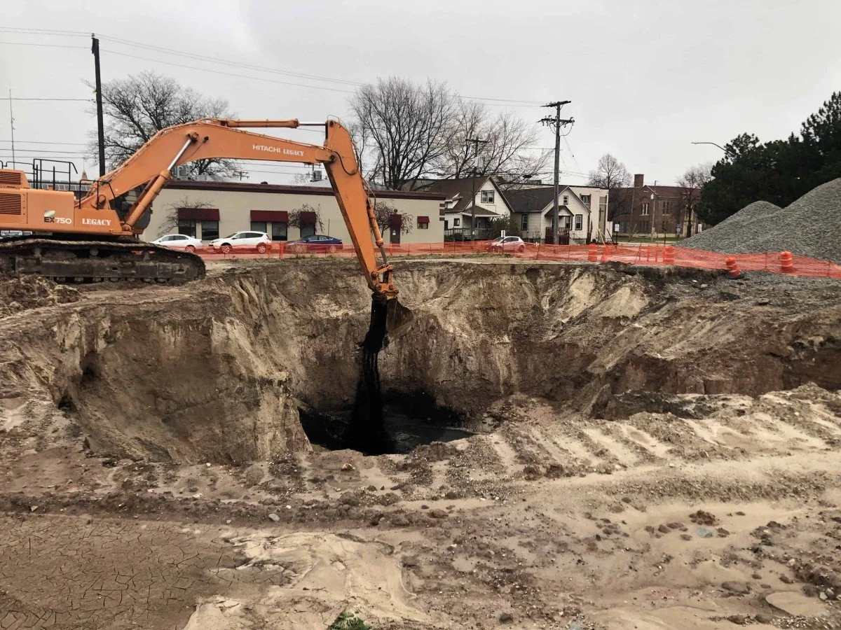 An excavator digging a large hole at a construction site, with residential houses and a parking lot visible in the background.