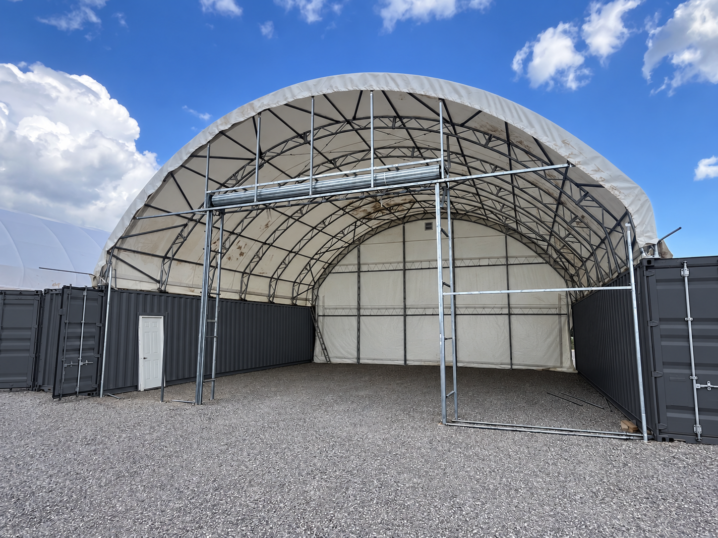 Large, open agricultural or industrial tent structure with metal frame, partially enclosed with dark and white panels, against a bright blue sky with clouds.