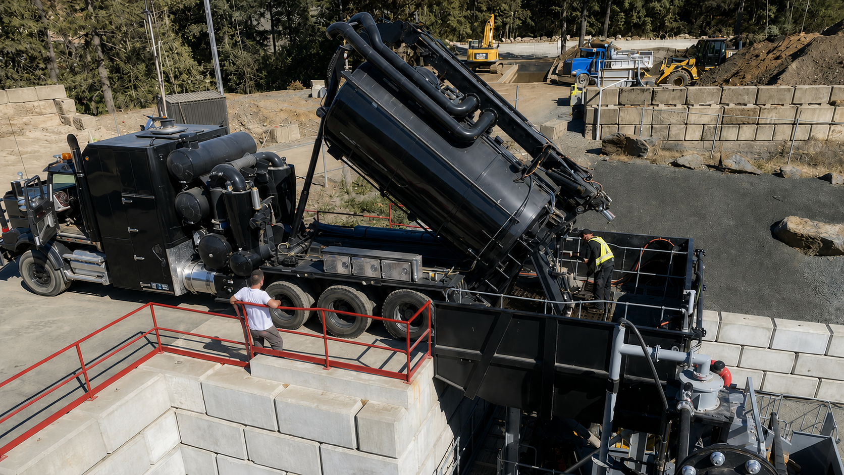A large black vacuum truck at a construction site with workers nearby. The truck's tank is tilted and connected to a system for removing or processing debris. Construction equipment and materials are visible in the background.