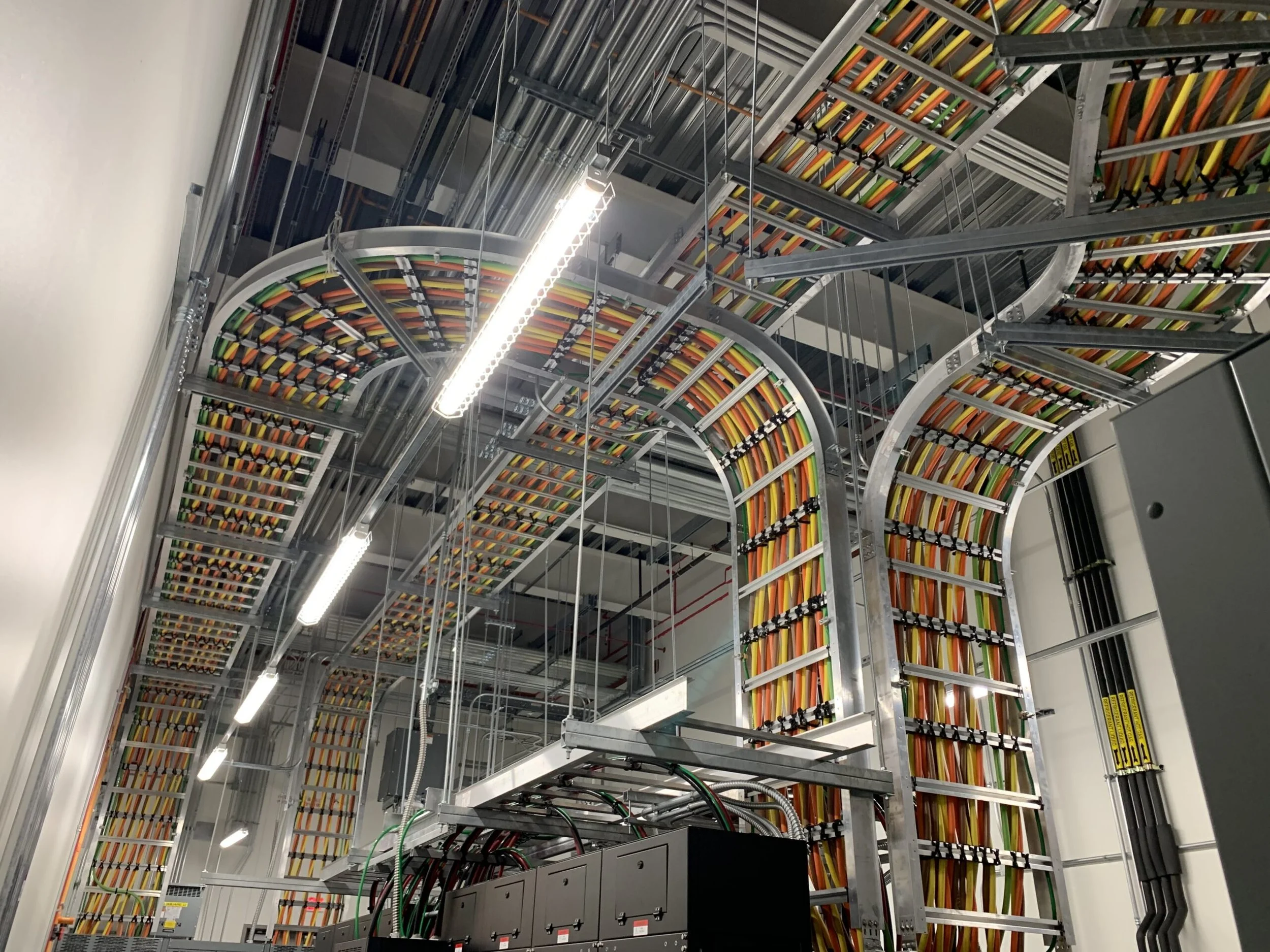 Inside a data center with cable management infrastructure, organized with multicolored cables routed through curved metal trays and conduit channels, illuminated by fluorescent lighting.