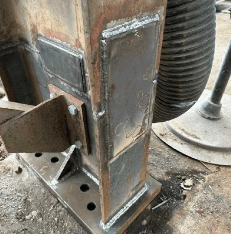 Close-up of a welded metal structure with bolts, on a concrete surface, with a flexible black duct and a metal pole nearby.