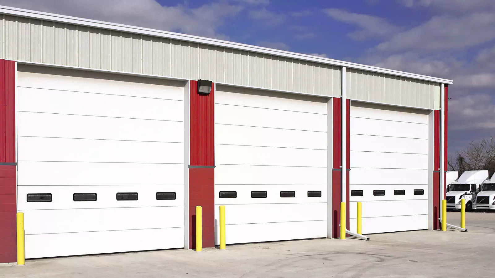 Three large white warehouse garage doors with black windows, red metal columns, and yellow safety bollards in front, with trucks parked to the right and a partly cloudy sky above.