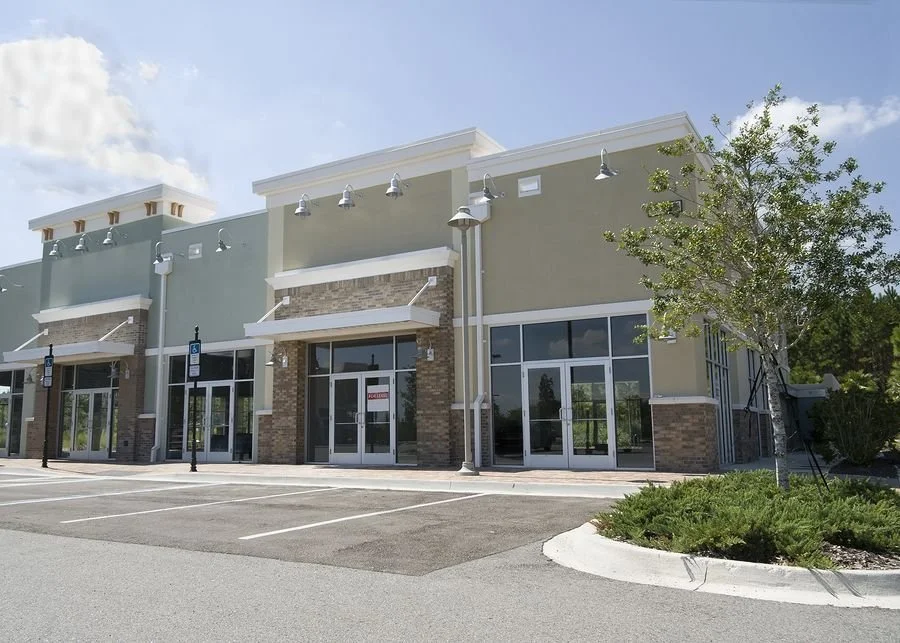 Newly constructed retail storefront with large glass windows and doors, parking lot with designated handicapped spaces, a tree and some greenery on the side.