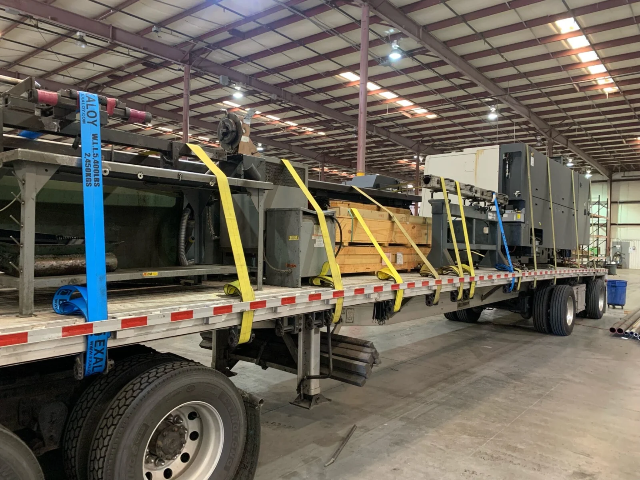 Large industrial machine secured on a flatbed truck inside a warehouse, with yellow and blue straps holding it down, and pipes and metal parts nearby.