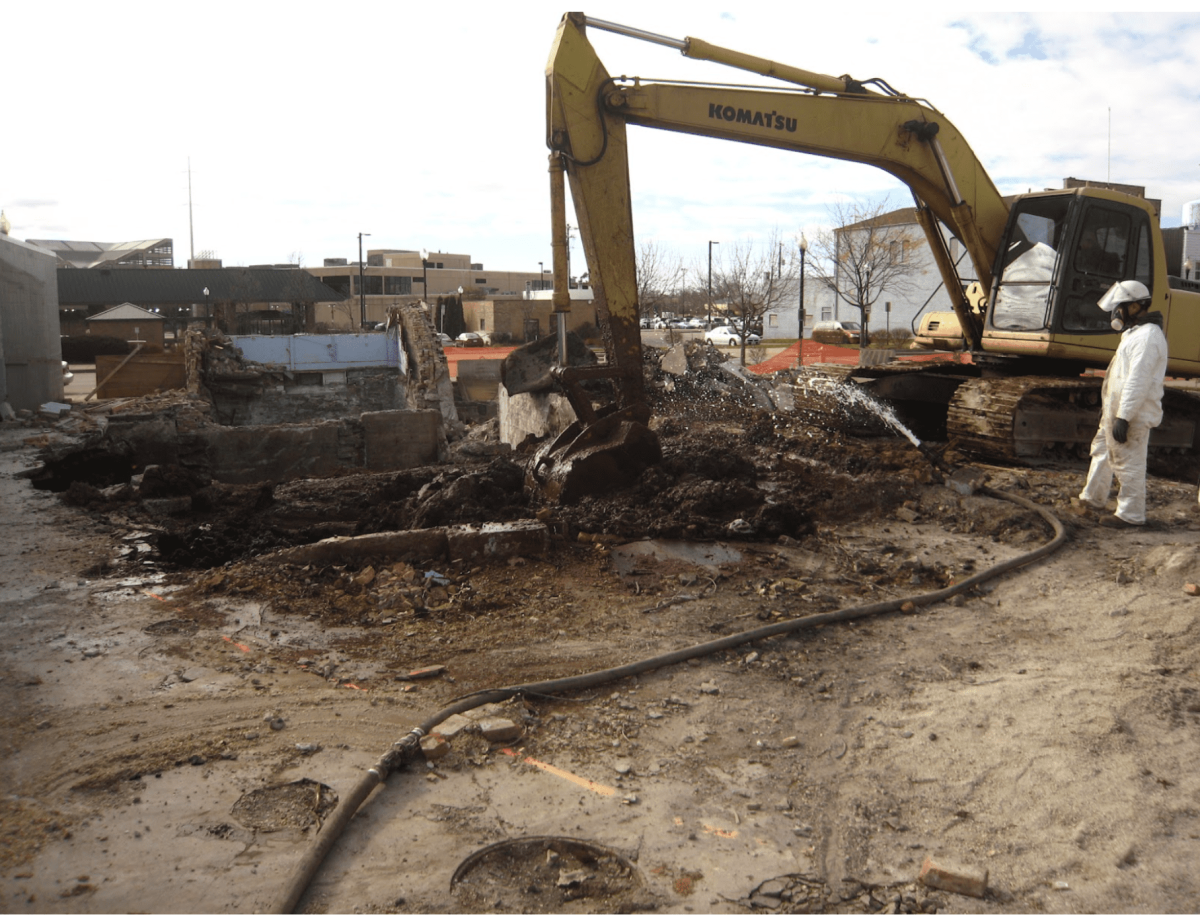 Construction site with a yellow Komatsu excavator demolishing a building, worker in white protective suit and helmet, and debris on the ground.