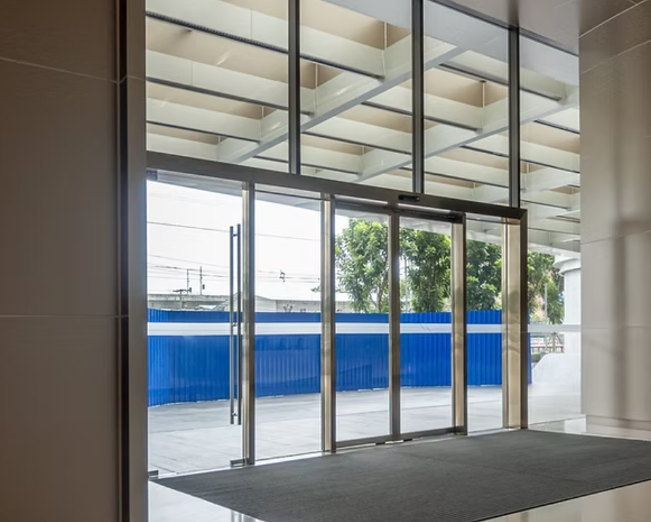 View of glass double doors leading outside to a patio or sidewalk with a blue fence, trees, and power lines visible beyond.