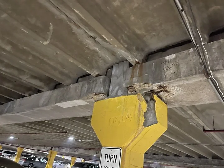 Concrete support beam in a parking garage with damaged yellow protective padding, exposed reinforcement, and painted markings.