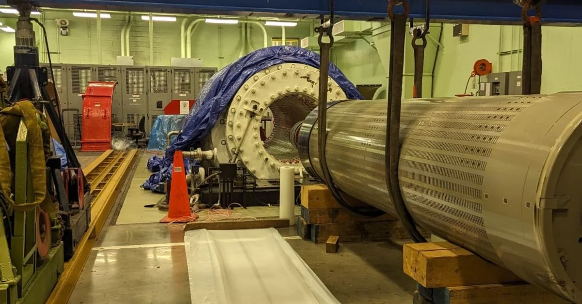 Large industrial machine or component undergoing maintenance or assembly inside a facility, with lifting straps, equipment, and safety cones surrounding it.
