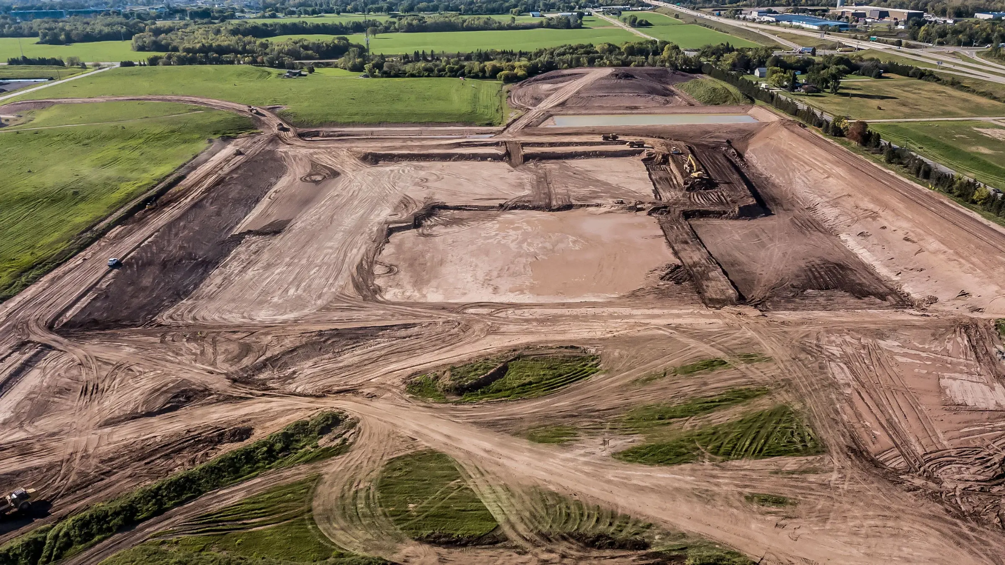 Aerial view of a large construction site with cleared and leveled land, multiple dirt roads, and construction equipment, surrounded by green fields and water bodies.
