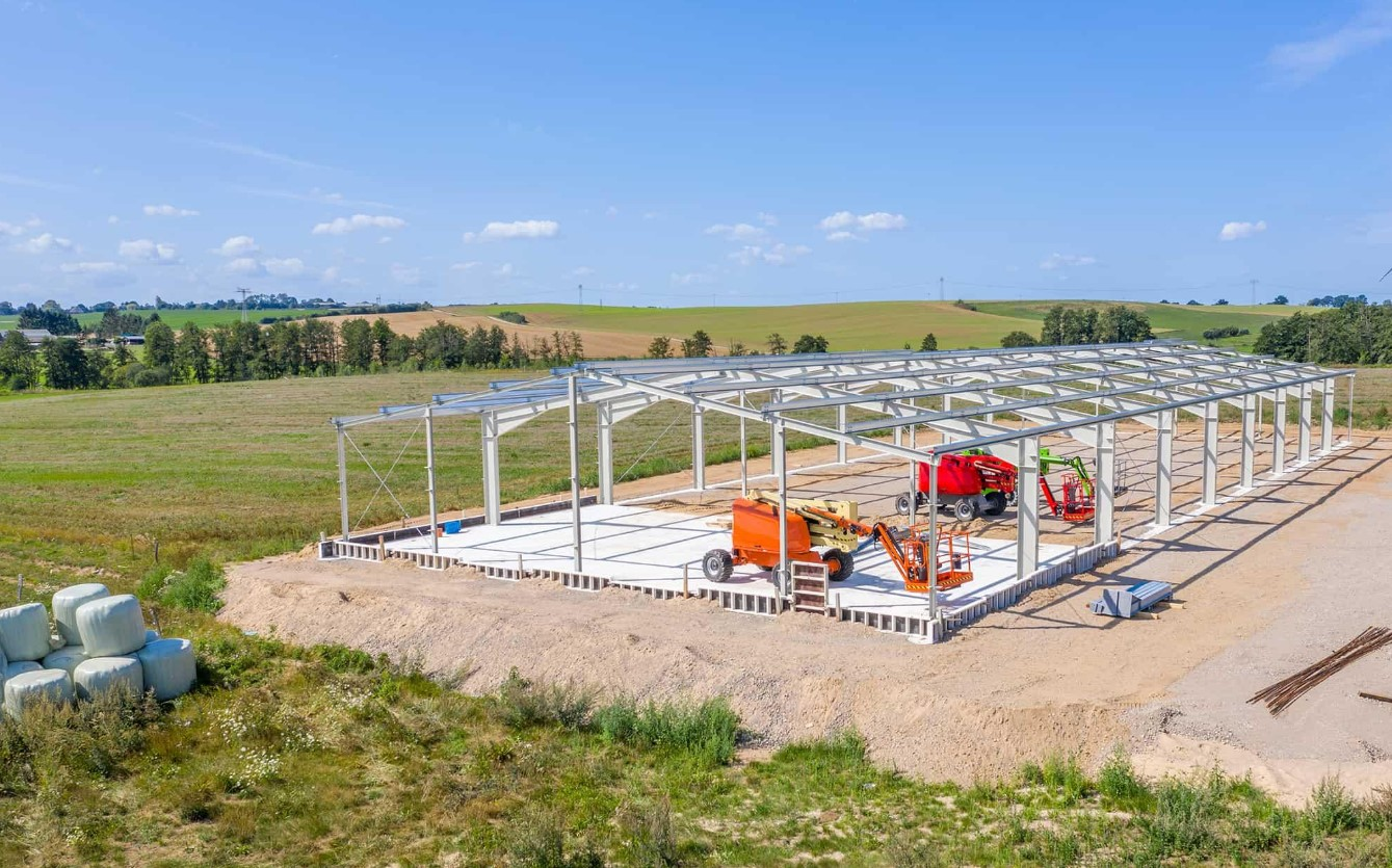 Construction site with a metal framework for a building, surrounded by green fields under a blue sky, with construction equipment inside the structure.