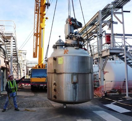 Industrial site with a worker observing a large cylindrical container being lowered by a crane
