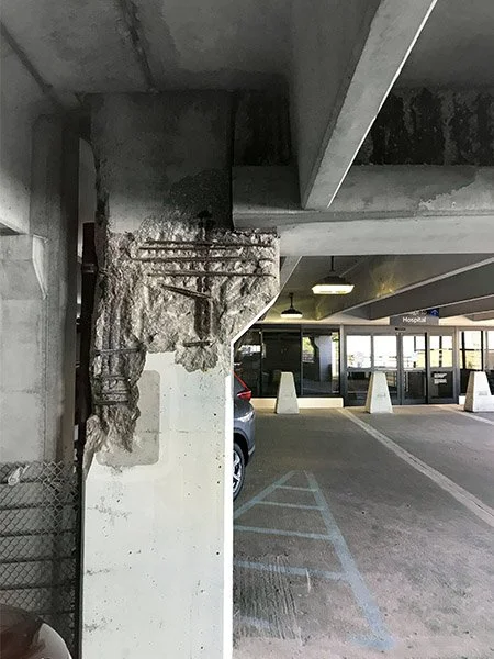A parking garage with exposed concrete beams and a damaged concrete support column with rebar.