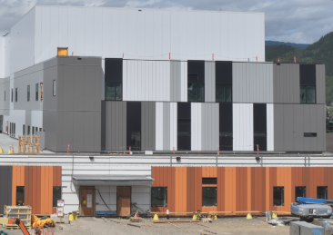 Under construction building with gray, black, white, and wooden exterior panels, construction equipment, and vehicles in front.