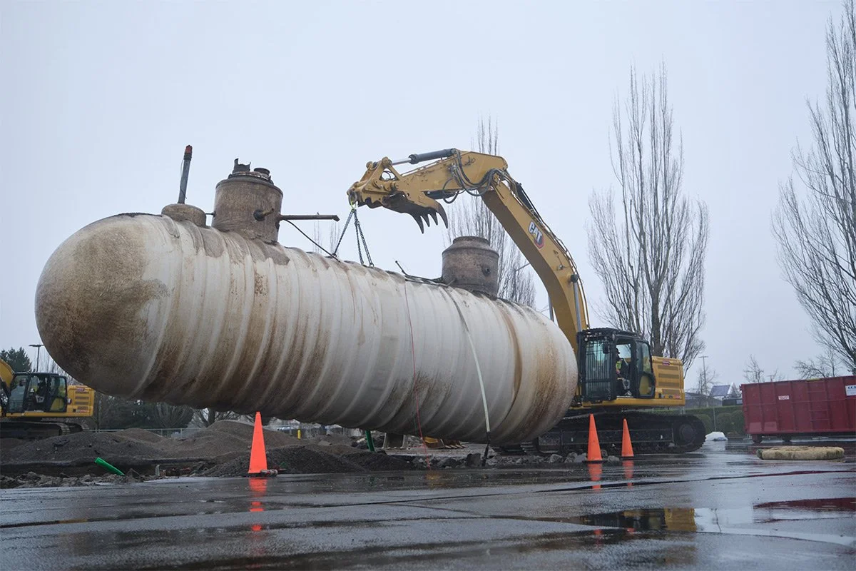 A large, rusty industrial tank is being lifted by a yellow Caterpillar excavator at a construction site on a rainy day, with orange cones and bare trees in the background.