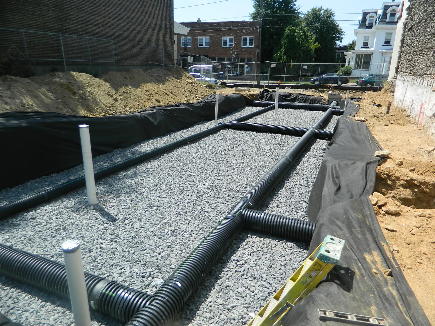Construction site with gravel foundation, black flexible pipes, and white pipes for plumbing or electrical work, surrounded by black fabric liner and wooden ladder.