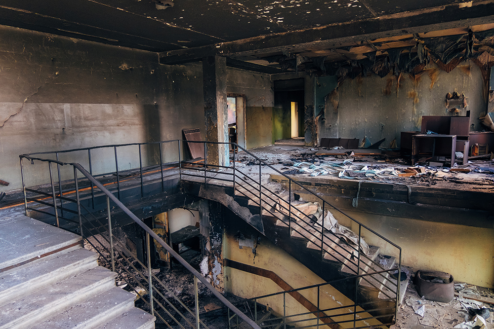 Interior of a burned down building with damaged walls, debris, and charred furniture, with a metal staircase and handrail.