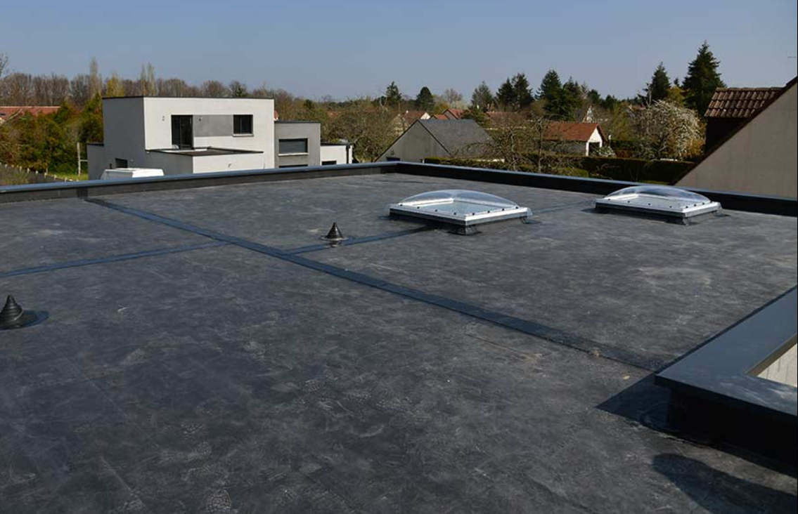 View of a flat rooftop with two skylights and a ventilation pipe, overlooking a neighborhood with modern and traditional houses, trees, and clear blue sky.