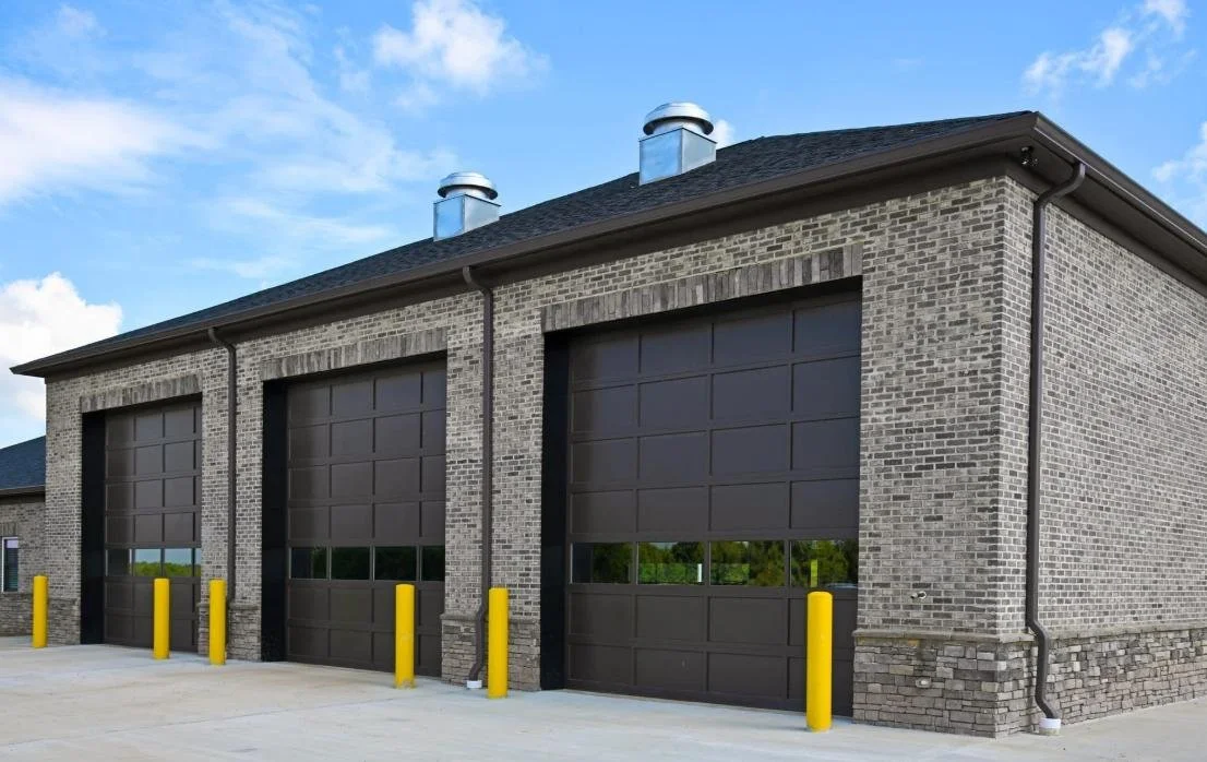 Three large black industrial garage doors on a brick building with yellow safety bollards in front, under a blue sky.