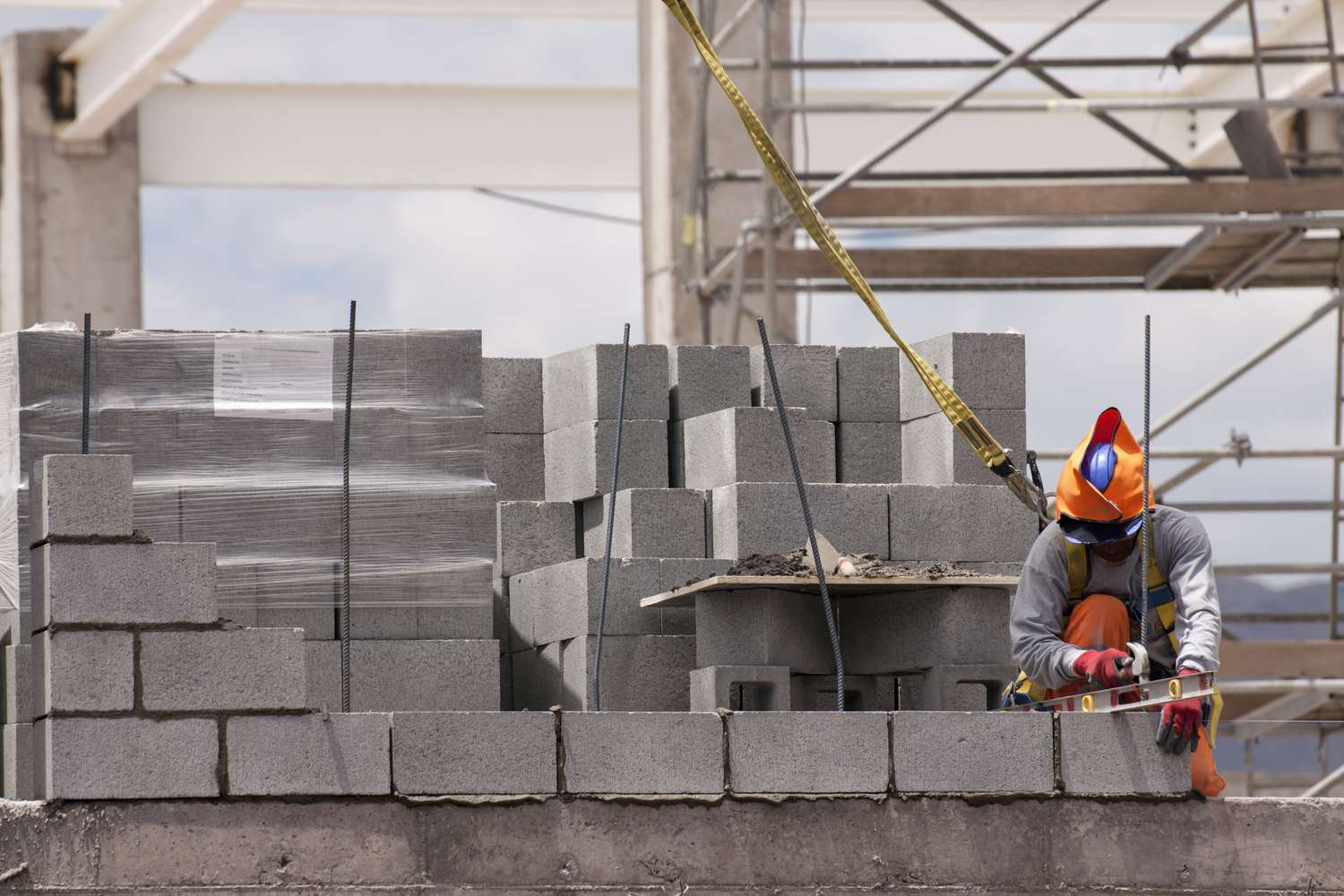 Construction worker wearing a helmet and safety gear, working with a leveling tool on a brick wall at a building site with scaffolding in the background.