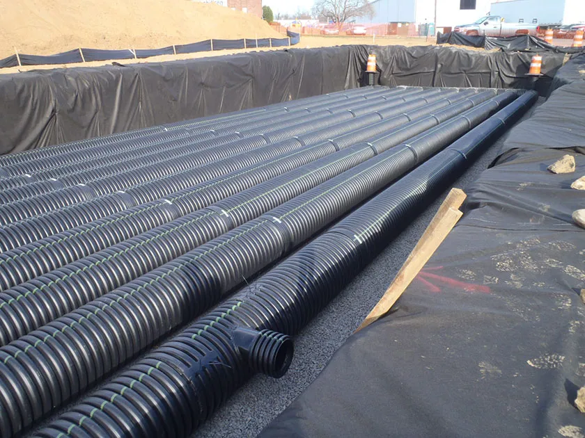 Construction site with black corrugated pipes laid in a trench, lined with black fabric, with orange safety barrels in the background.
