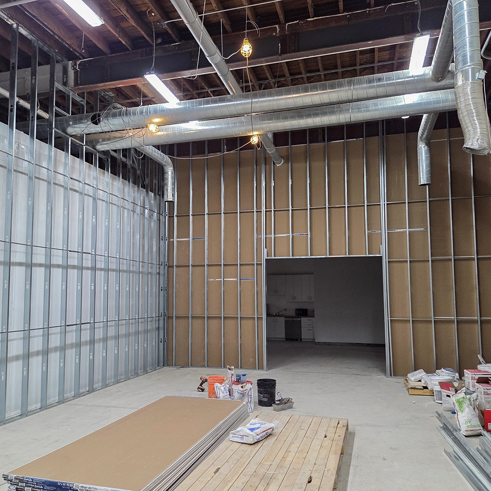 Interior of a building under construction with exposed metal framing, ventilation ducts, and construction materials on the floor.
