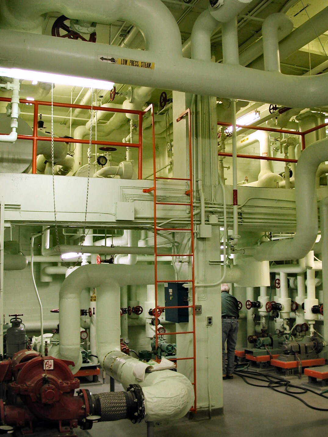 An industrial mechanical room with white pipes, red safety railings, and a worker in the background.