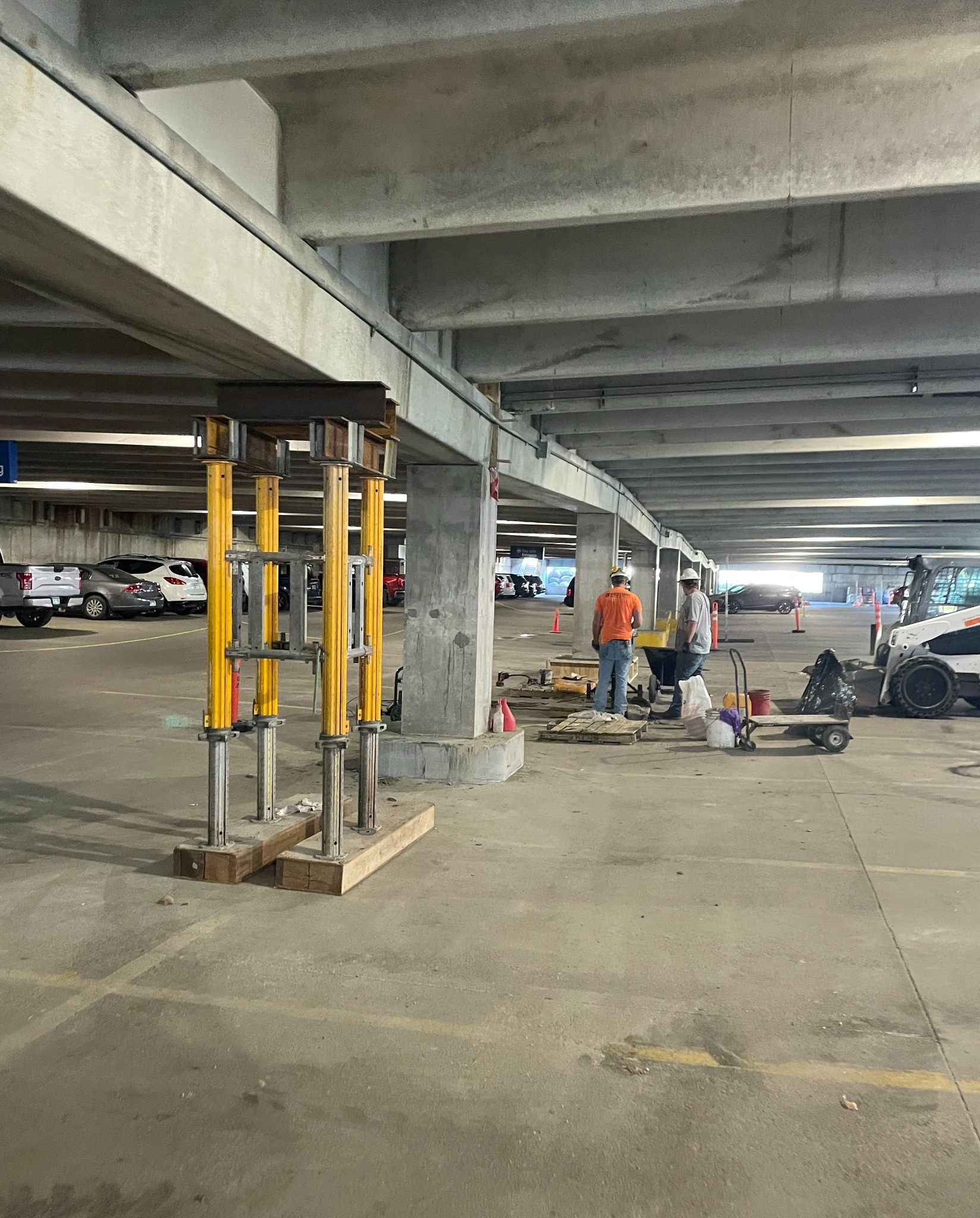 Construction workers working in an underground parking garage with construction equipment and cars parked nearby.