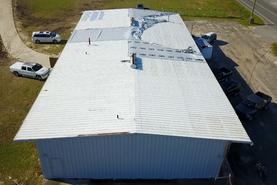 Aerial view of a large metal building with a white roof, some areas of damage, and a line of parked vehicles nearby.