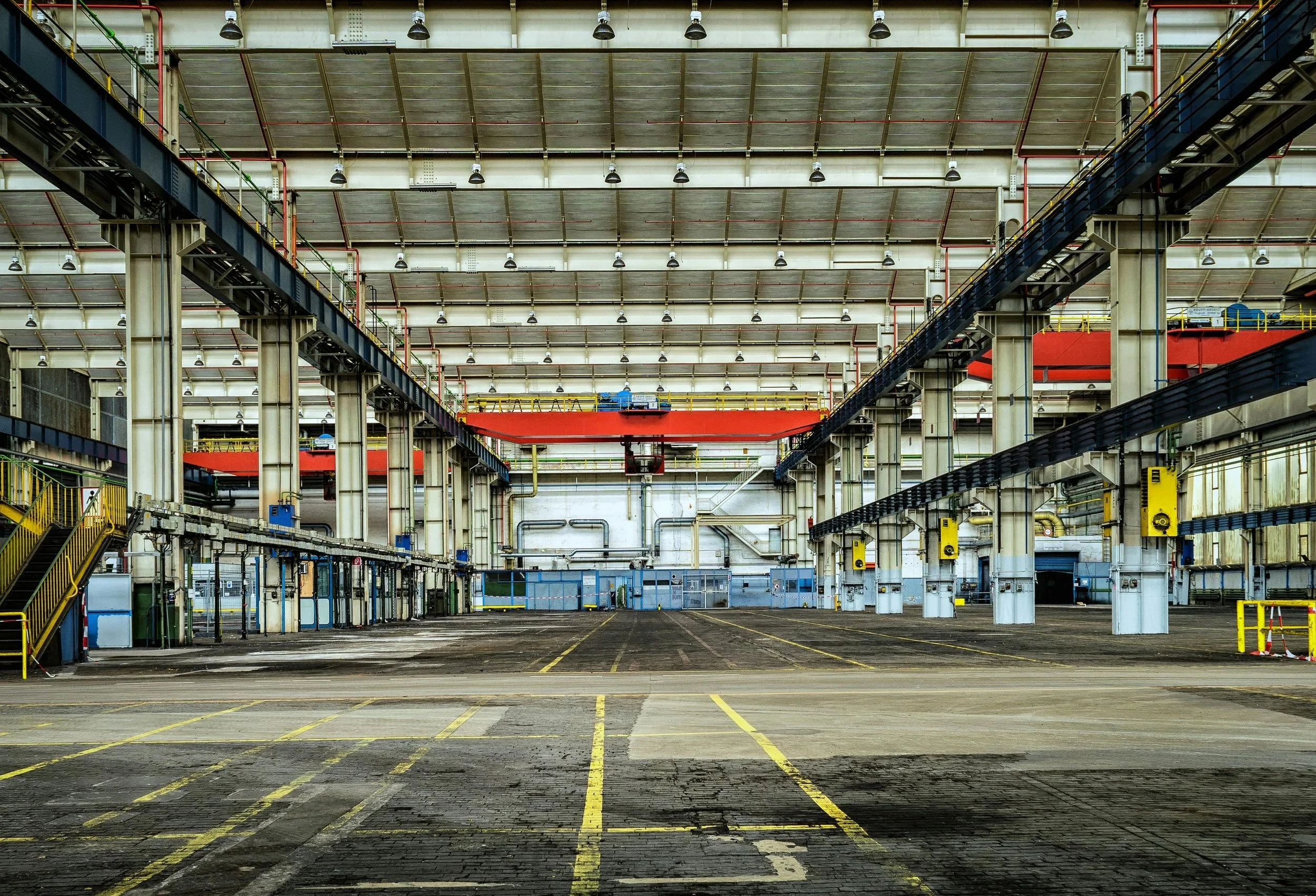 Empty industrial warehouse with yellow lines on the floor, large overhead cranes, and blue and yellow structural supports.