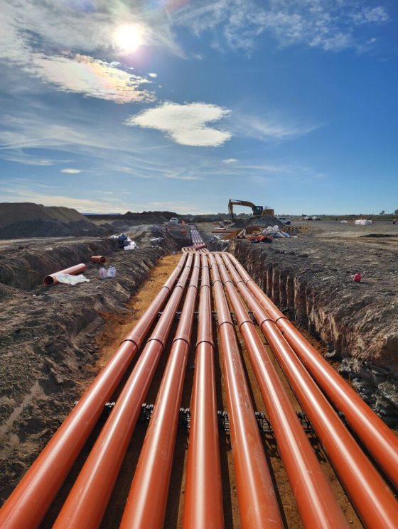 Construction site with orange pipes laid in a trench, with construction equipment and supplies in the background under a partly cloudy sky.