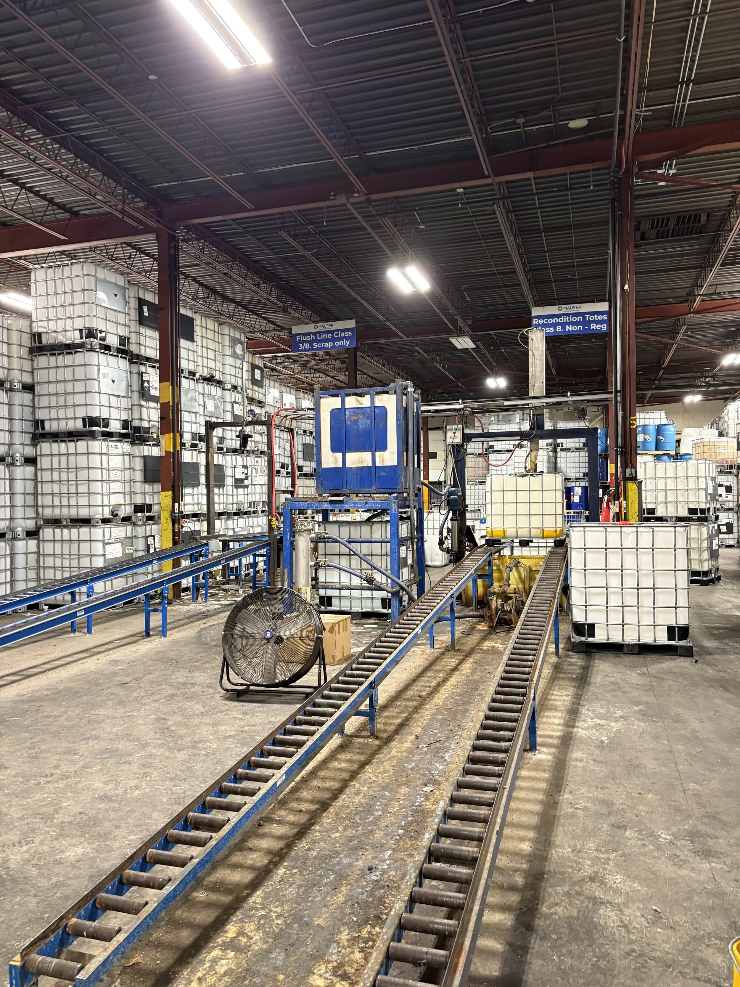Industrial warehouse with large white plastic containers stacked on metal shelves, conveyor belts, and reconditioning equipment. Signage indicates areas for flush line class and reconditioning totes.