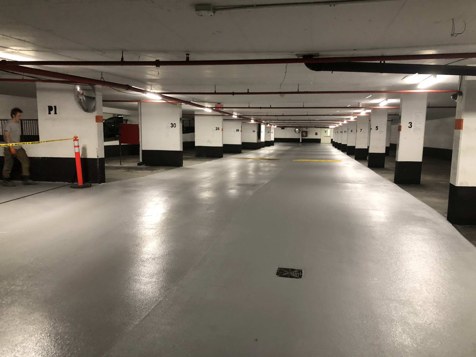 Empty underground parking garage with white pillars numbered 1 to 30, illuminated overhead fluorescent lights, and a man walking past a red cone on the left side.