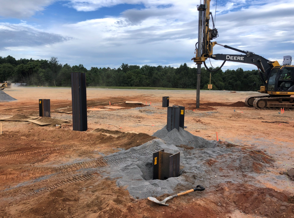 Construction site with steel pilings, a dirt ground, a dirt excavator, and a cloudy sky.