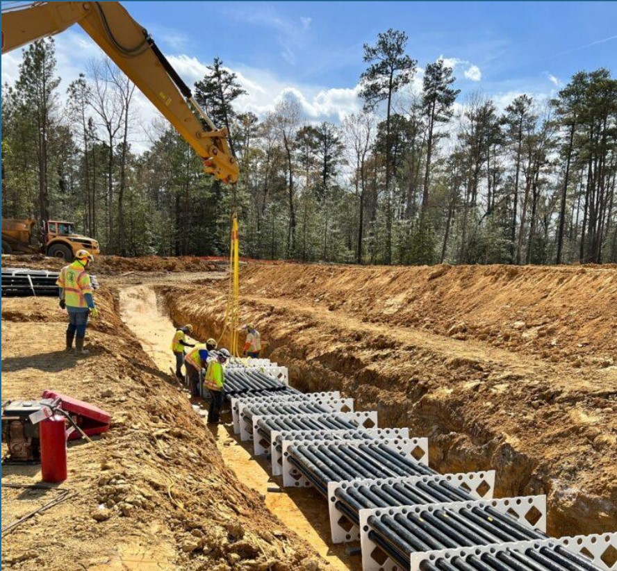 Construction workers in safety gear installing large pipes in a trench at a construction site, with trees and a blue sky in the background.