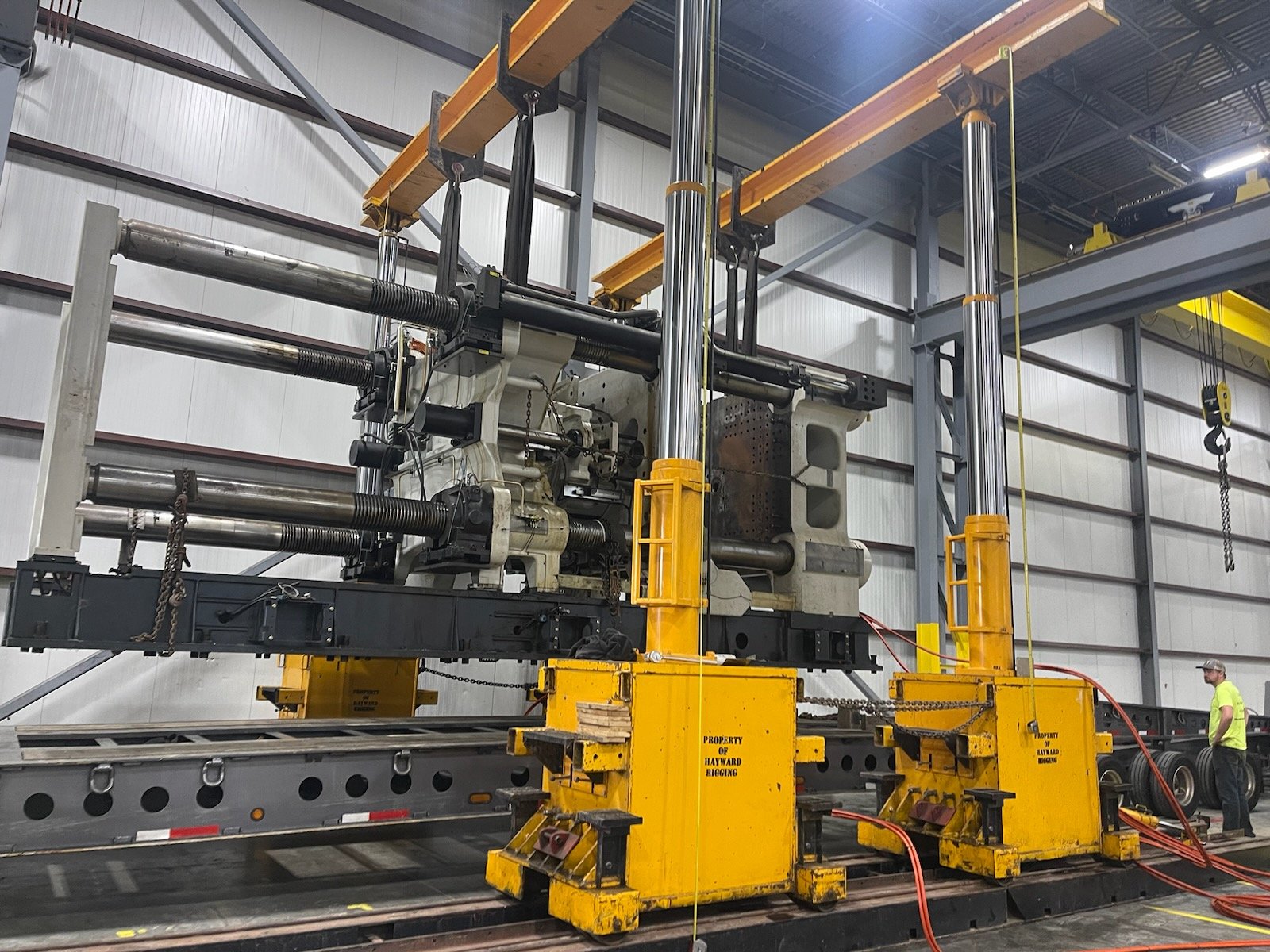Large industrial machine used for testing or manufacturing, situated on a trailer in a warehouse, with a worker standing nearby.