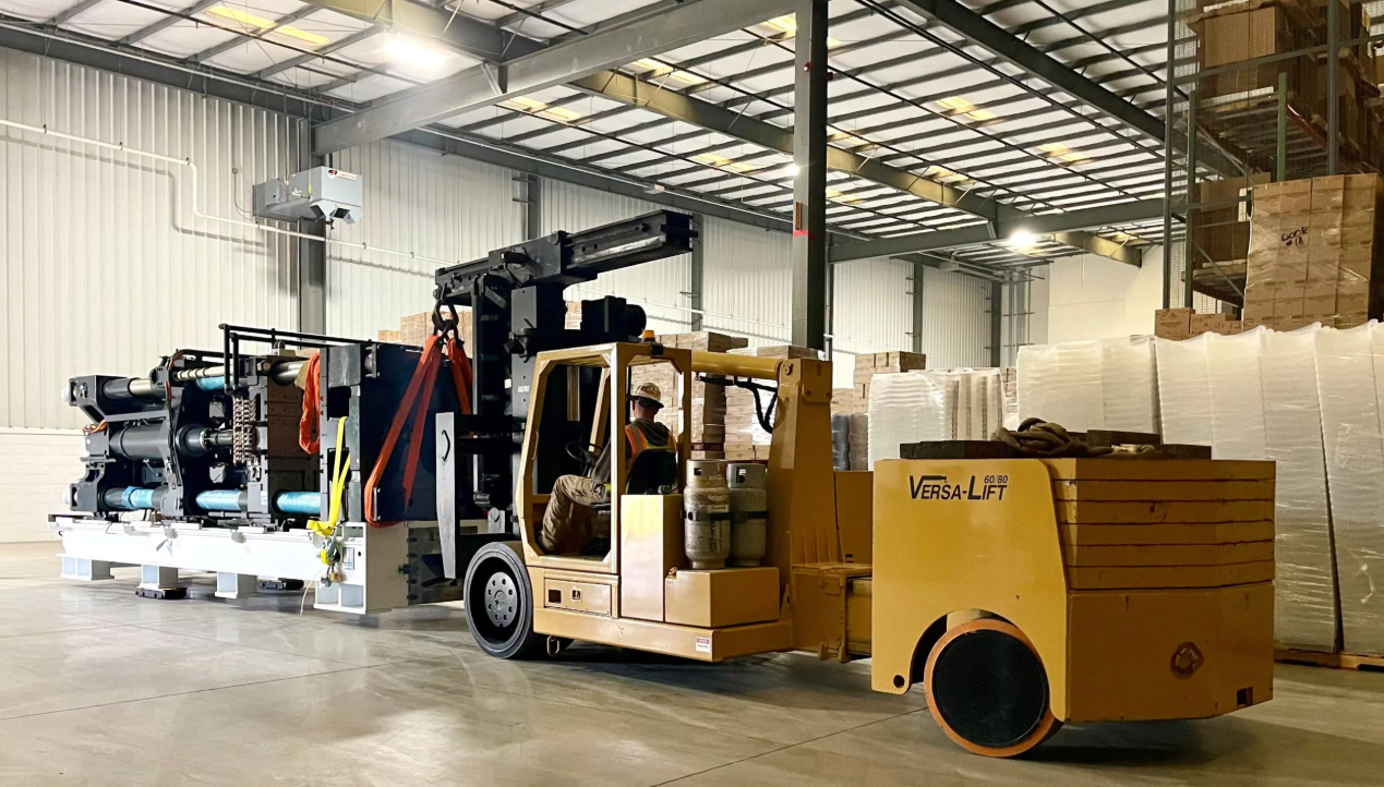 A forklift operator is moving stacks of boxes and pallets inside a warehouse using a yellow Versa-Lift forklift, with large rolls of plastic wrap and boxes on shelves in the background.