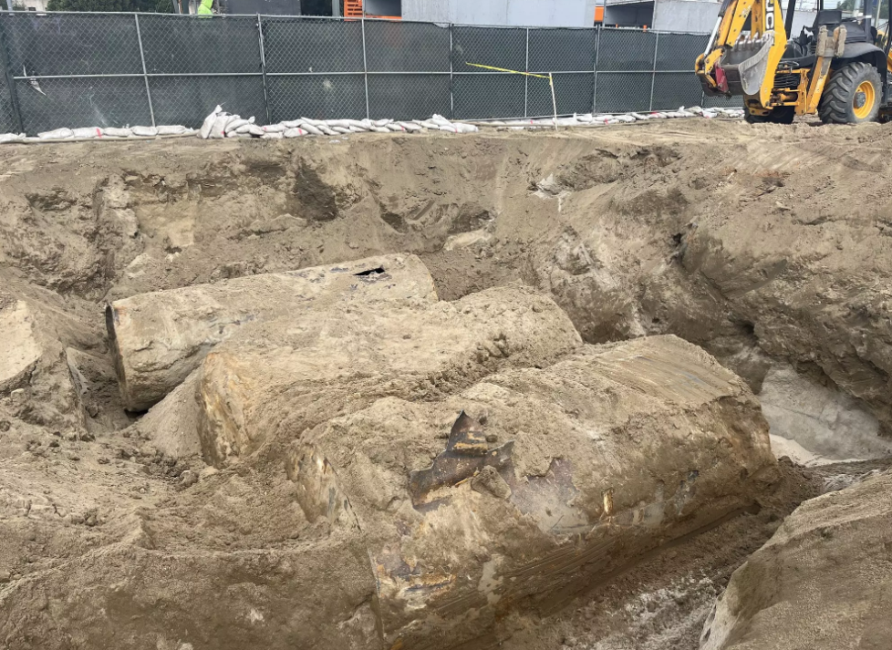 Excavation site with large buried remnants of an object, construction machinery, and a fenced-off area in the background.