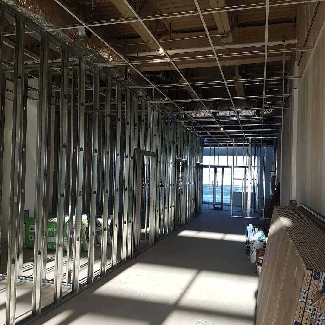 Interior of a building under construction with metal wall framing, ceiling grid framework, and construction materials on the floor.