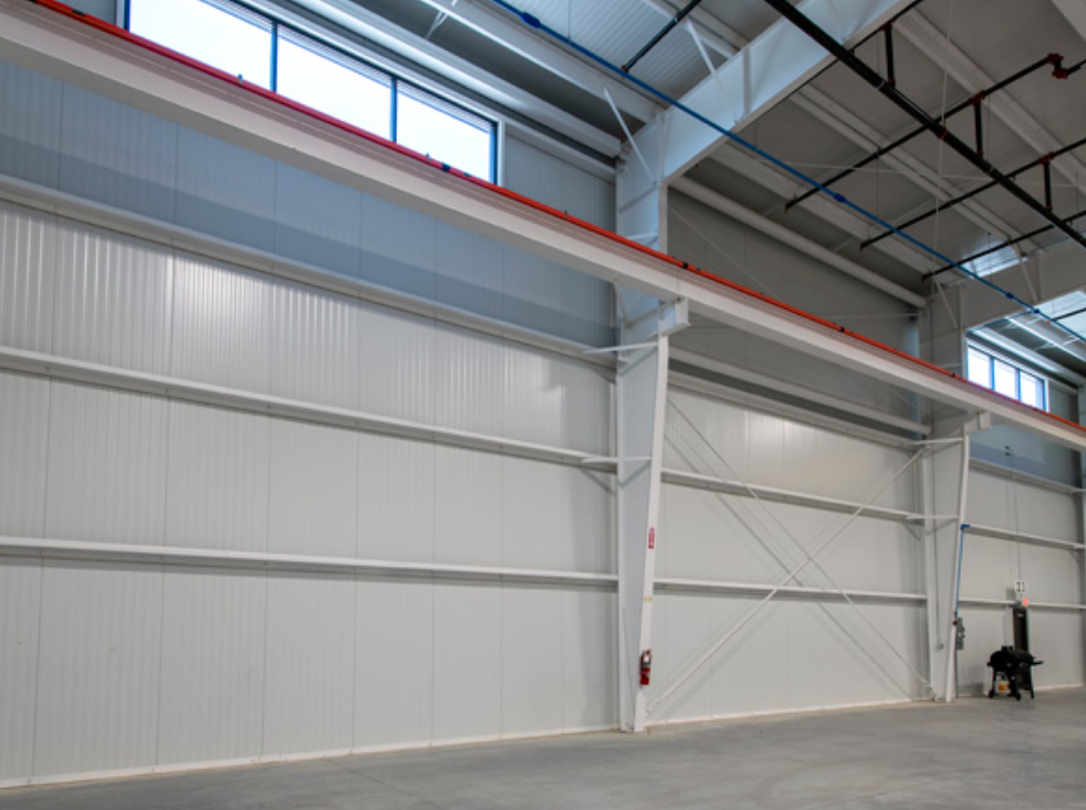 Interior of a large industrial warehouse with high ceilings, metal walls, and large windows.