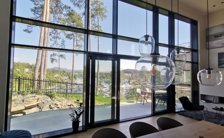 Modern living room with large glass windows and doors, overlooking a lake and trees, with hanging glass pendant lights and a balcony with outdoor furniture.