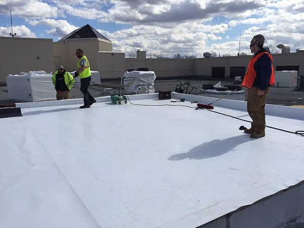 Workers in safety vests applying white coating on a flat rooftop, surrounded by tools and supplies, under a partly cloudy sky.
