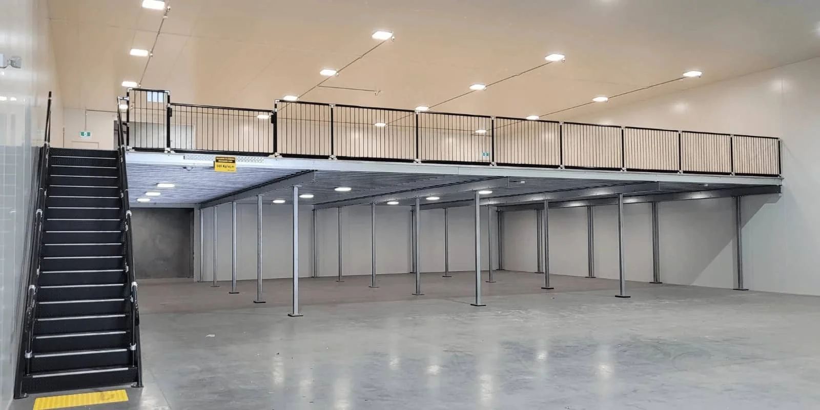 Empty retail store or warehouse with a mezzanine level, metal support columns, concrete floor, and a staircase leading up to the second floor.