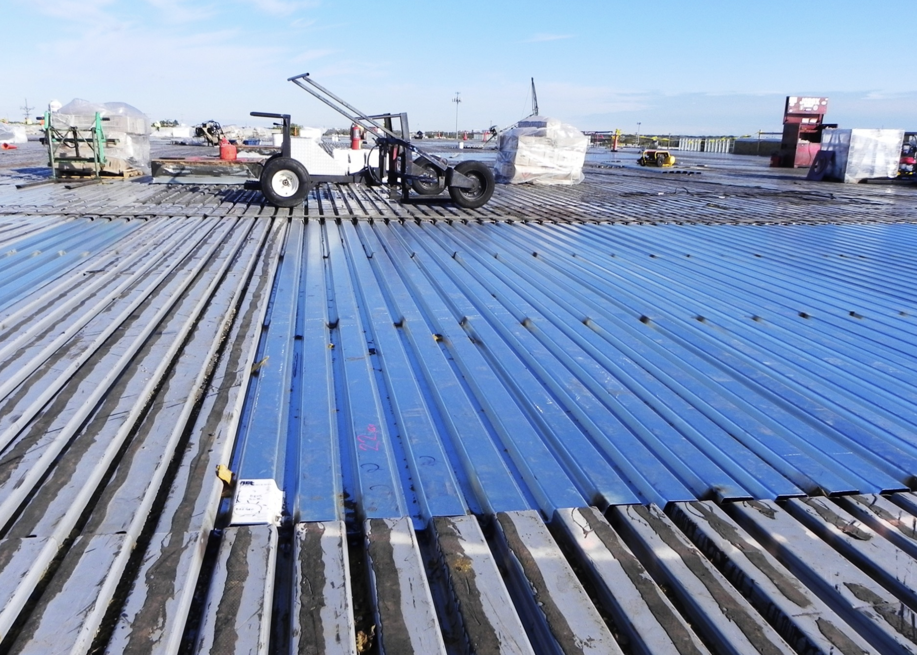 Construction site with metal roofing panels being installed, with equipment and boxes in the background, under a blue sky.