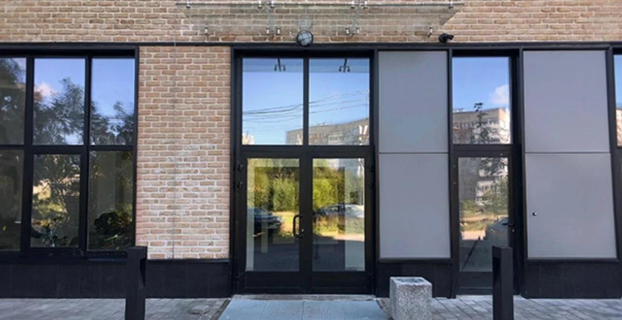 Modern building entrance with black-framed glass doors and large windows, brick wall, and gray panels, with a concrete block and black posts outside.