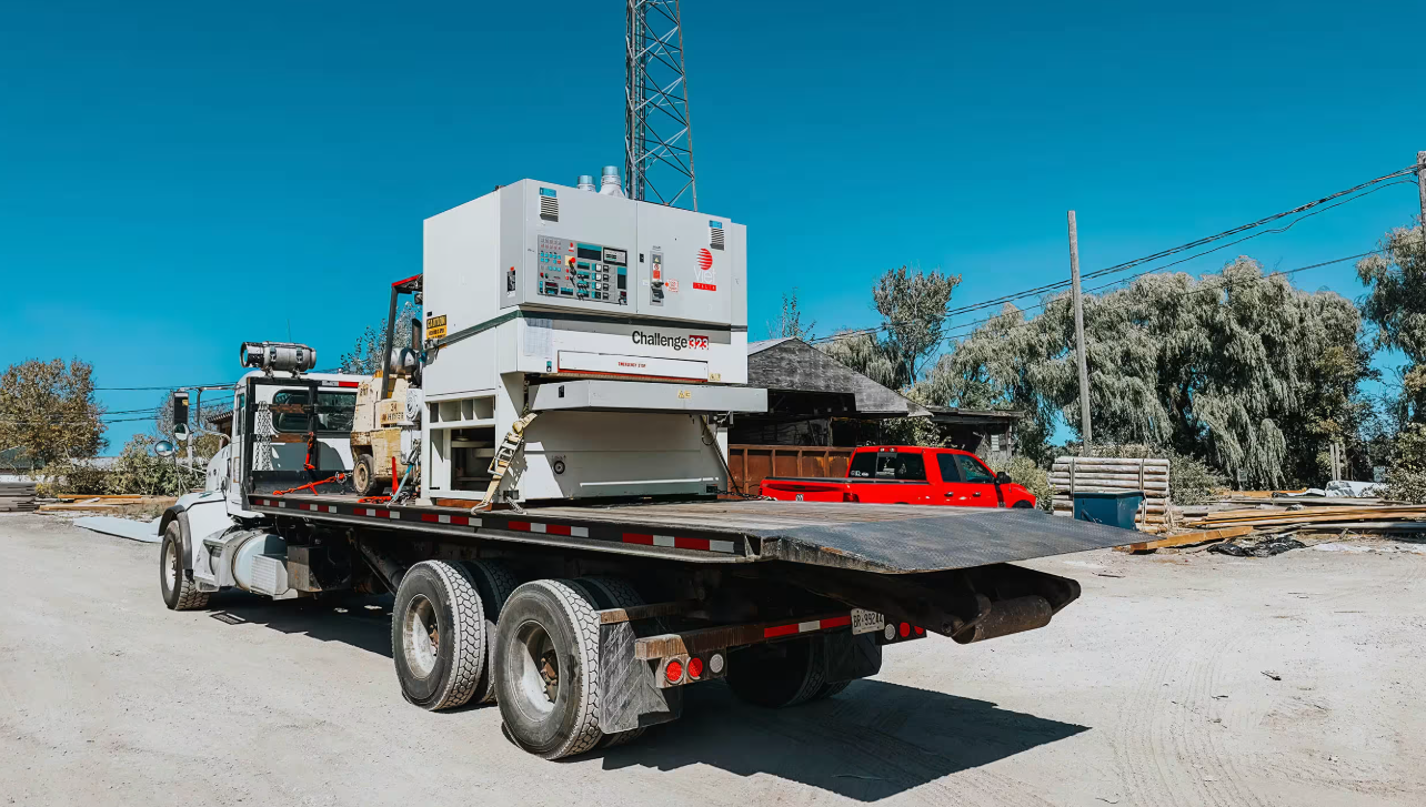 A flatbed truck carrying a large commercial generator and other equipment parked on a gravel lot.