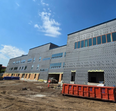 Construction site with a large, modern building under construction, dirt ground, and construction equipment under a blue sky with some clouds.