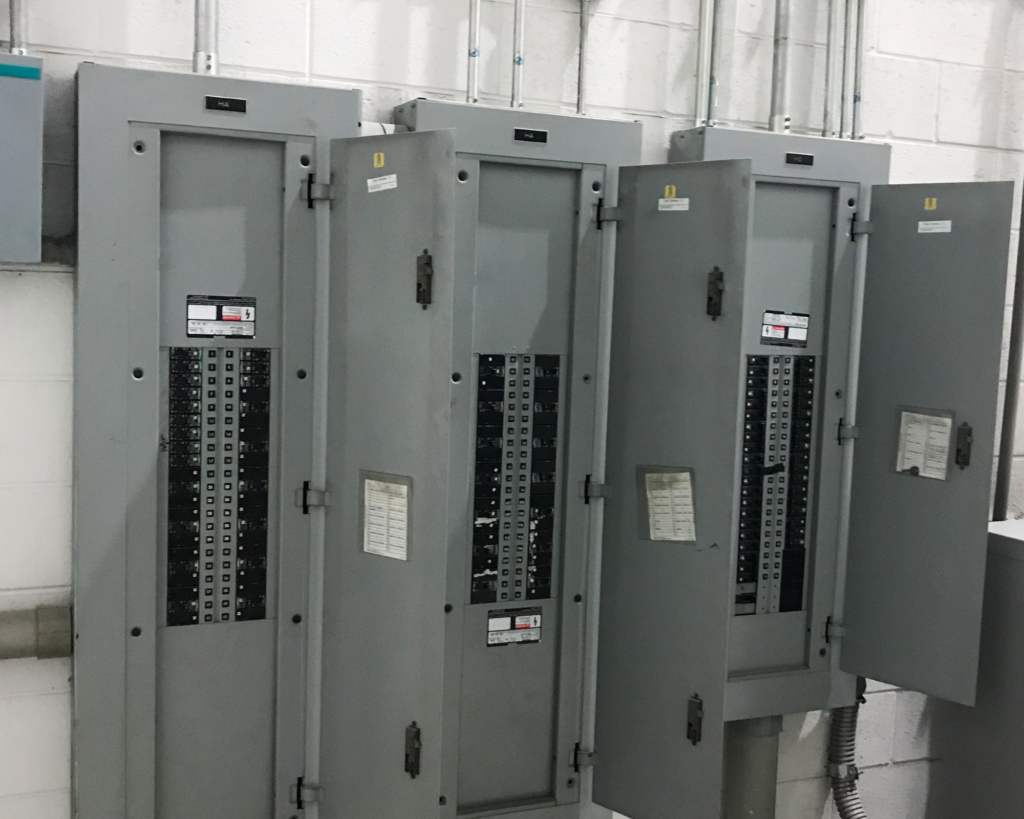 Four gray electrical breaker panels mounted on a white brick wall in an industrial setting.