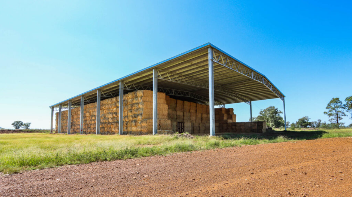 Large open shelter with metal framework filled with stacked hay bales on a farm under a blue sky.