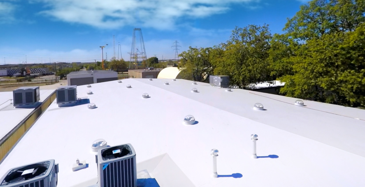 View of a white commercial building roof with multiple air conditioning units and vents, surrounded by trees and utility poles under a partly cloudy sky.